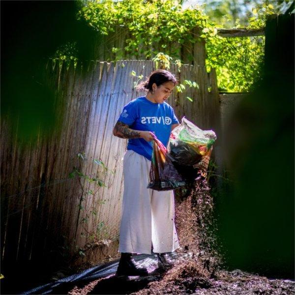 Alex-Mari Ford works on spreading mulch in a yard. She stands next to a fence and holds a bag of mulch.